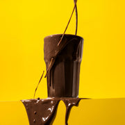 Black protein powder bag with a purple label next to a glass of chocolate protein powder on a light background